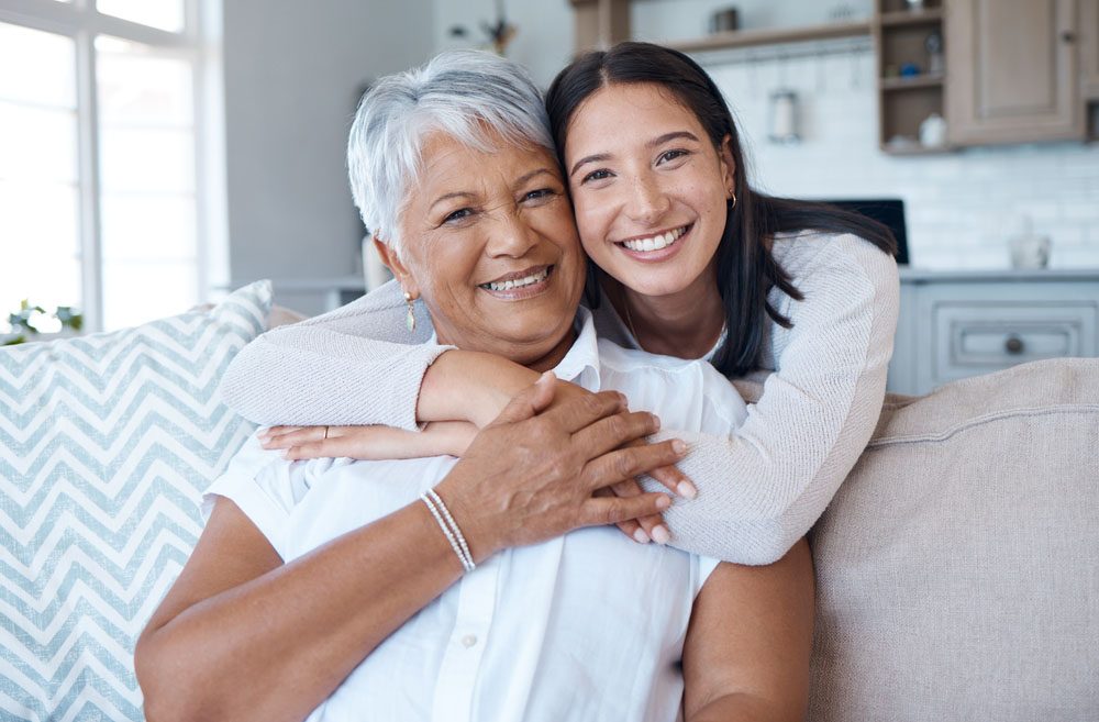 Young woman hugs an older woman from behind as they sit on a couch, both smiling warmly in a bright living room.