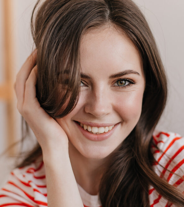 Close-up portrait of a smiling young woman with brown hair resting her head on her hand.