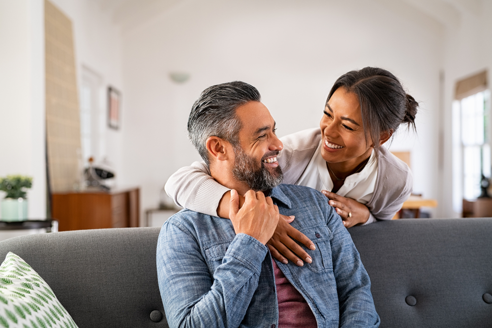 Smiling couple sharing a warm embrace on a couch in a bright living room.
