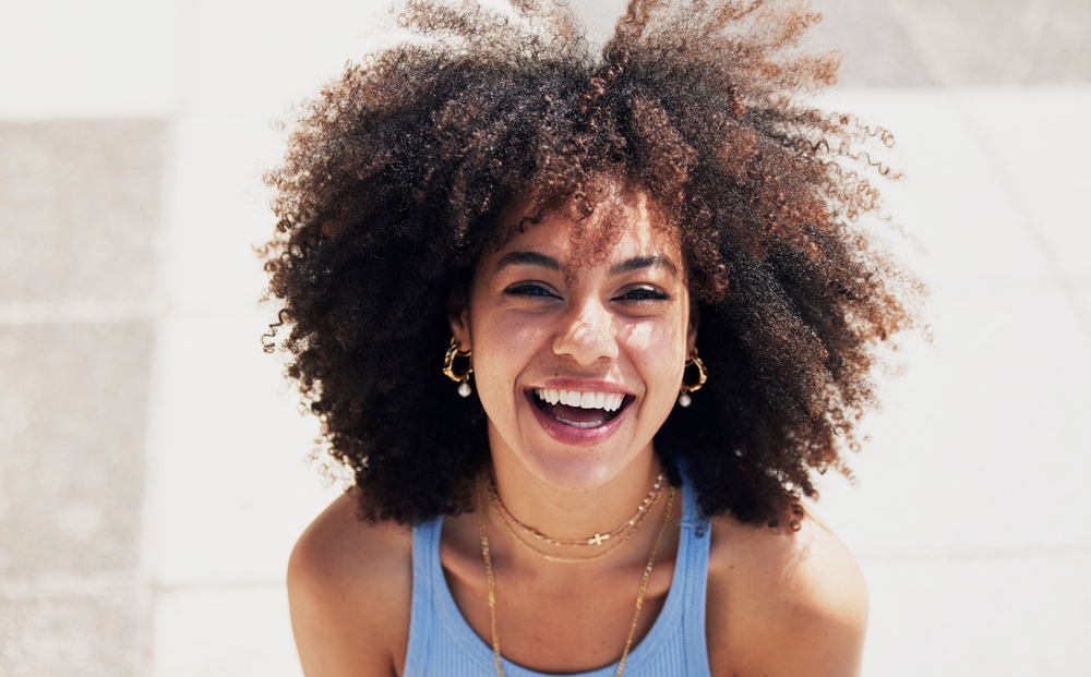 Smiling woman with curly hair laughs outdoors in bright sunlight, wearing a blue top and gold jewelry.