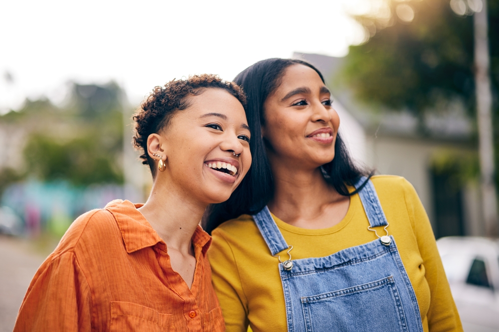 Two young women smiling together outdoors, looking off into the distance.