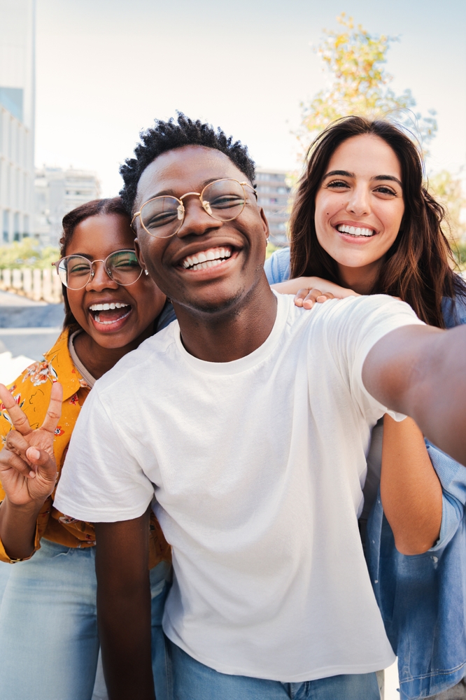 Three friends smiling and taking a selfie outdoors, with one person holding up a peace sign.