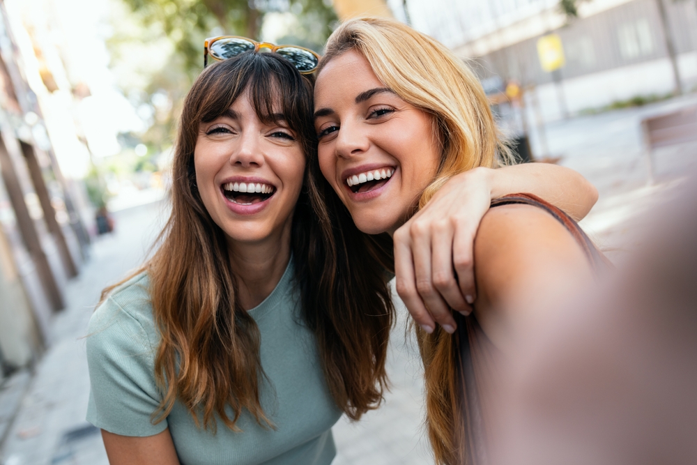 Two young women smiling and taking a selfie together outdoors on a city street.