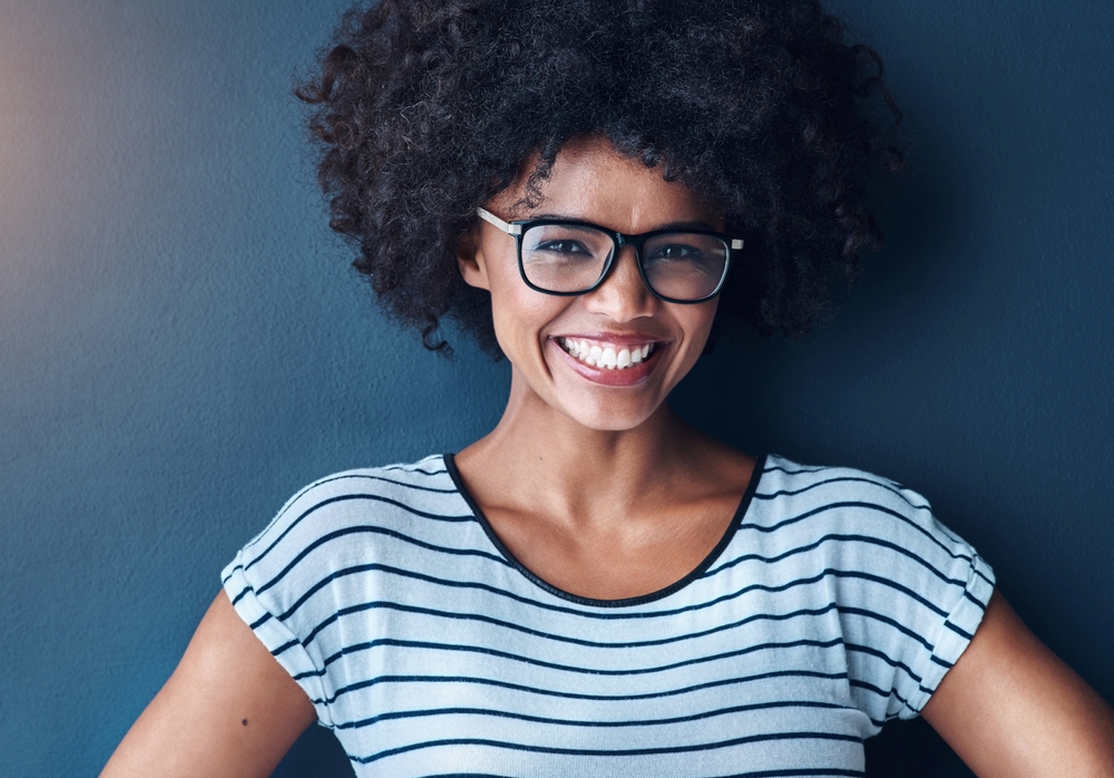Smiling woman with curly hair and glasses wears a striped shirt against a dark blue background.