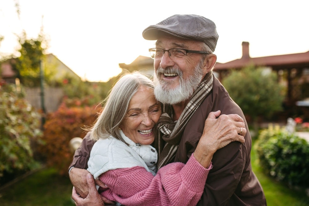 Elderly couple smiling and hugging outdoors in a garden setting during warm sunlight.