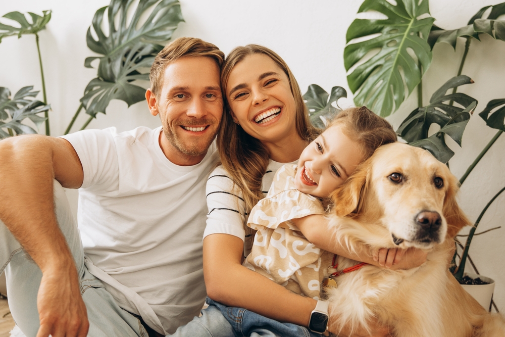 Smiling family sitting together with their young child and golden retriever in a cozy home surrounded by green plants.