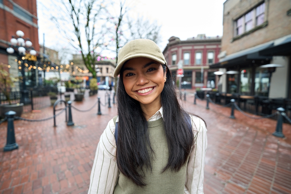 Smiling young woman wearing a cap stands on a brick street in an outdoor urban setting.