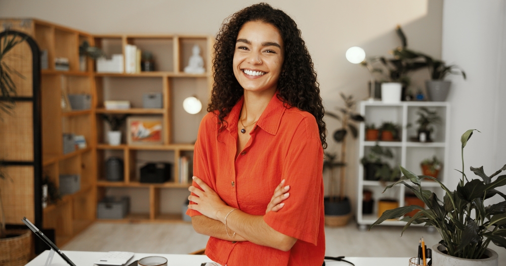 Smiling woman with curly hair stands with arms crossed in a modern office with plants and shelves in the background.