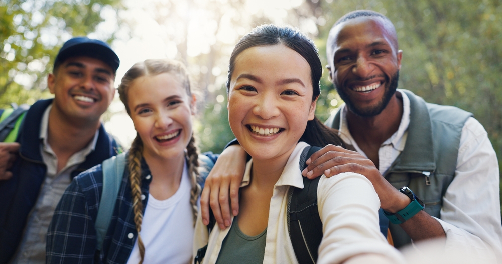 Group of friends smiling and taking a selfie outdoors while hiking with backpacks.
