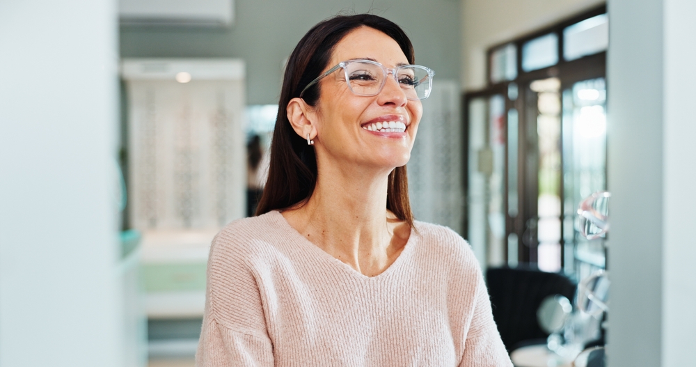 Smiling woman wearing clear eyeglasses sits in a bright modern office or clinic setting.