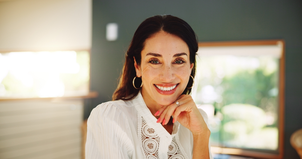 Smiling woman with dark hair and hoop earrings rests her chin on her hand in a softly lit indoor setting.