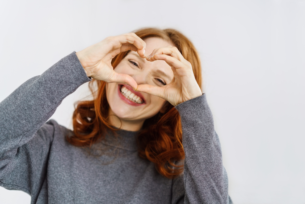 Smiling red-haired woman makes a heart shape with her hands in front of her face against a light background.